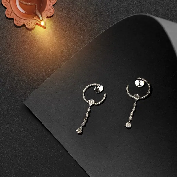 Diamond hoop drops on folded black card, a diya glowing at the corner.