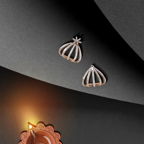 Rose-gold cage earrings on black paper, with a diya burning below.